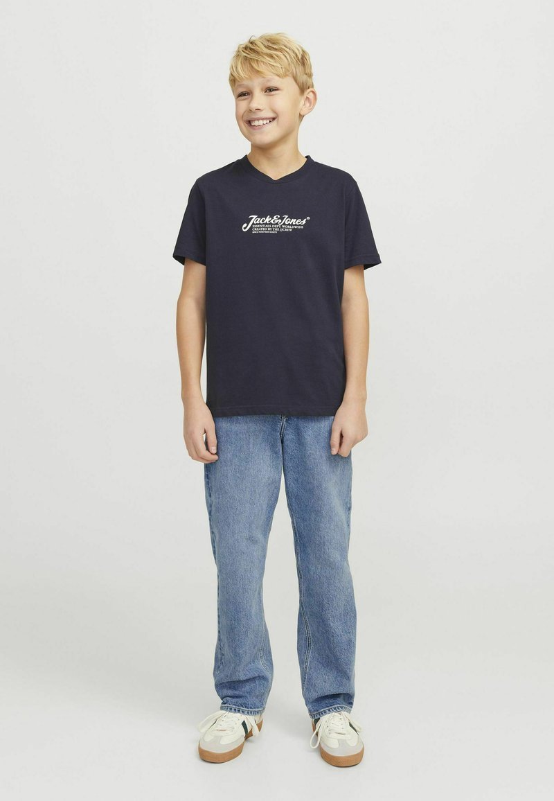 Camiseta de algodón azul marino con escote en V y logo blanco, combinada con jeans de mezclilla azul claro y zapatillas crema con acentos verdes.
