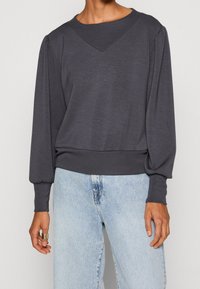 Donkergrijze pullovertrui met een V-hals, lange pofmouwen en geribbelde zoom. Gemaakt van zachte stof, gecombineerd met lichtblauwe jeans.