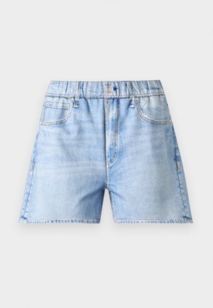Sort kvinde stående med hånden på hoften iført hvid gennemsigtig overdel, lyseblå denim shorts og sorte sandaler med gyldent hjerte-detalje.