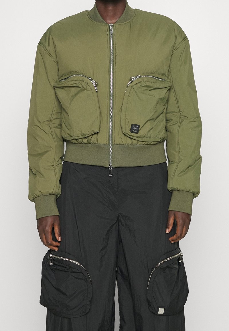 Haut du corps portant une veste bomber vert olive avec deux grandes poches zippées, associée à un pantalon noir avec de grandes poches zippées sur les cuisses.