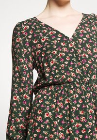 Robe fleurie avec un fond vert, ornée de fleurs roses et blanches, décolleté en V, manches longues et taille froncée pour un ajustement fluide.