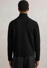 Juoda megztinis su aukšta apykakle ir ilgomis rankovėmis. Turi raukšlėtą tekstūrą ir tiesų apatinį kraštą. Minimalistinis dizainas.