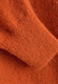 Primer piano di un morbido e soffice tessuto di maglia arancione, con polsini a costine ben in vista, che mostra la trama e il motivo della lavorazione a maglia.