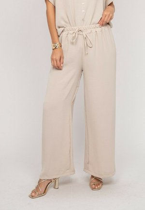 Femme portant un pantalon beige à jambes larges avec une taille élastique et un cordon de serrage, associé à des talons dorés à brides et un bracelet manchette doré.