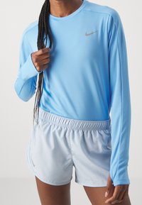 Person med långt flätat hår som bär en ljusblå långärmad top från Nike och matchande sportshorts, som håller i en fläta med ena handen.