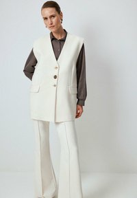 Chaleco blanco sin mangas con escote en V, cuatro botones y dos bolsillos frontales. Combinado con pantalones acampanados blancos y una camisa de botones oscura por debajo.