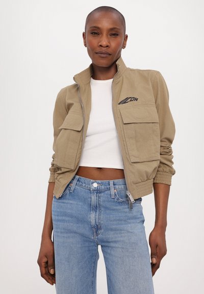 Khaki värvi bomber-jakk, millel on ees lukuga sulgur, kaks rinnataskut ja elastsed varrukad. Kantud valge crop topi ja helesiniste teksade peal.