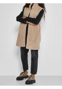 Béžová vesta z umělého beránka s vysokým límcem, zapínáním na patentky a bočními kapsami, kombinovaná s černým oblečením a masivními hnědými botami.