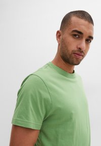 T-shirt a maniche corte con scollo rotondo in cotone verde chiaro. Texture liscia con dettagli di cucitura lungo le maniche e il colletto.