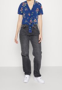 Person iført en blå blomsteret t-shirt med korte ærmer, højtaljede falmede sorte jeans og hvide sneakers, der står mod en ensfarvet baggrund.