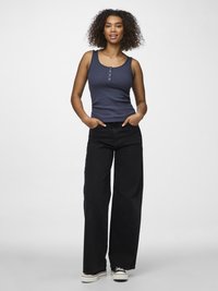 Donna con capelli ricci che indossa una canotta blu navy a costine, pantaloni neri a gamba larga e scarpe da ginnastica bianche e nere, in piedi con le mani nelle tasche.