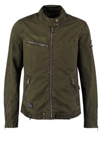 Olivengrüne Jacke aus leichtem Stoff. Sie hat einen hohen Kragen, mehrere mit Reißverschluss versehene Taschen und einen Reißverschluss zur Schließung. Inklusive Steppdetails.