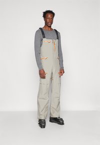 Ljusgrå overall med orange dragkedjor och detaljer, justerbara axelremmar och flera fickor. Ett par svarta skidpjäxor fulländar outfiten.
