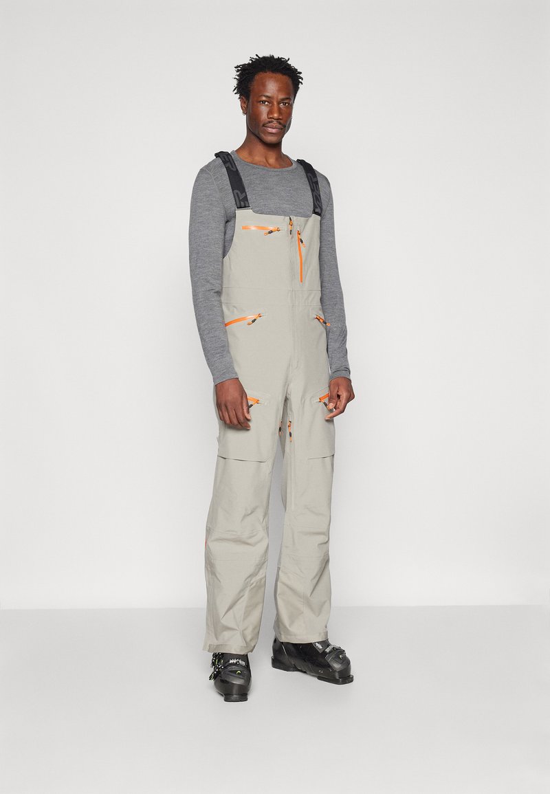 Ljusgrå overall med orange dragkedjor och detaljer, justerbara axelremmar och flera fickor. Ett par svarta skidpjäxor fulländar outfiten.