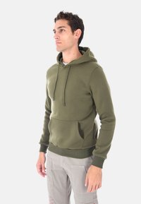 Uomo in piedi che indossa un ragazzo verde oliva con tasca anteriore e pantaloni cargo grigi chiari, che guarda a sinistra su uno sfondo bianco.