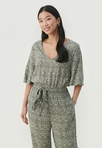 Mlada žena z dolgimi temnimi lasmi, oblečena v zeleno-belo vzorčasto kombinezon s pasom, se smeje in ima roke v žepih ob beli podlagi.