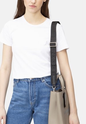 Borsa tote grigia con tracolla nera, caratterizzata da una texture liscia e branding in rilievo. Indossata con una t-shirt bianca e jeans blu.