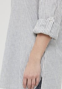 Camicia a righe con linee blu e bianche, caratterizzata da maniche arrotolate con bottone, uno spacco laterale e una texture di tessuto leggero.