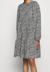 Robe à imprimé floral avec des manches longues, col rond et jupe à volants. Présente un fond marine avec des fleurs blanches et bleu clair. Tissu en coton.