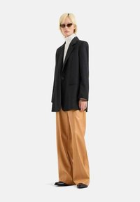 Blazer nero su una maglia a collo alto bianca, abbinato a pantaloni larghi color cammello e scarpe nere, su uno sfondo neutro.