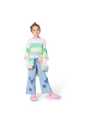 Kolorowy sweter w paski w zielonym, różowym i niebieskim kolorze, jasnoniebieskie dżinsy z szerokimi nogawkami z wzorem serc oraz różowe chodaki z ozdobnymi detalami.