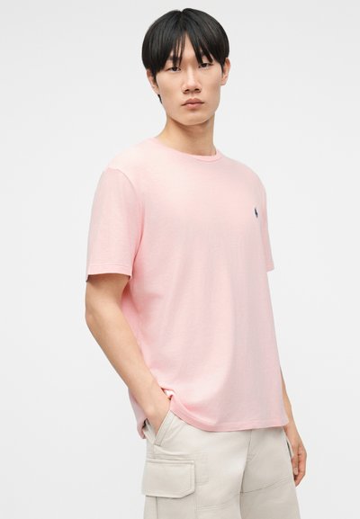 Νέος άνδρας με μαύρα μαλλιά που φοράει ένα ανοιχτό ροζ κοντομάνικο μπλουζάκι και μπεζ σορτς cargo, στέκεται με το ένα χέρι στην τσέπη.