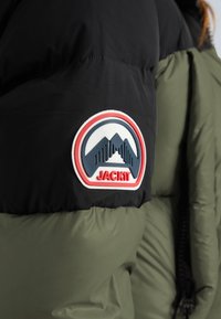 Patch with mountain logo and "JACKIT" text on the sleeve of a black and olive green puffer jacket.