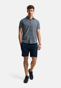 Camisa de manga corta, con botones, a rayas en azul marino y blanco, combinada con pantalones cortos azul marino y zapatillas oscuras. Tejido ligero y casual.
