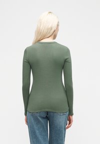 Γυναίκα με μακριά ξανθά μαλλιά, φορώντας στενή ριμπ πλεκτή πράσινη μπλούζα με μακριά μανίκια και μπλε τζιν, στέκεται γυρισμένη με την πλάτη, σε λευκό φόντο.