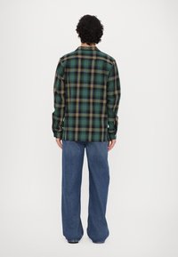 Grønn og svart rutete skjorte med gule detaljer, lange ermer, knappede mansjetter og en avslappet passform, kombinert med vide blå jeans.