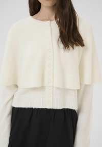 Fehér, réteges pulóver kerek nyakkal, bordázott mandzsettákkal és gombos zárással, puha, textúrált anyagból készült. Fekete nadrággal párosítva.