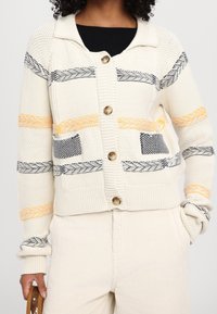 Kremowy sweter robiony na drutach z kołnierzem, w żółte, czarne i szare paski, z zapięciem na guziki i dwiema przednimi kieszeniami.