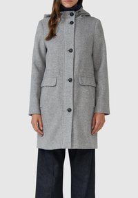 Grijze wollen jas met een capuchon, voorzien van een knoopsluiting, twee voorzakken en een textuurafwerking. Gecombineerd met donkere spijkerbroek.