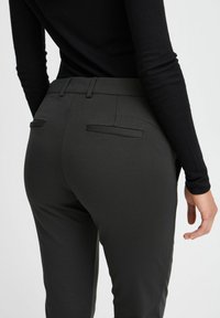 Pantaloni verde scuro su misura con una texture liscia, dotati di due tasche posteriori e un design slanciato. Abbinati a un top nero attillato.