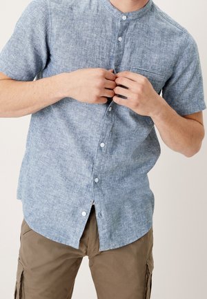 Person, die ein kurzärmeliges, hellblaues Chambray-Hemd über braunen Cargo-Hosen vor einem schlichten Hintergrund zuknöpft.