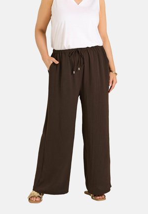 Frau trägt locker sitzende dunkelbraune weite Hosen mit Kordelzug in der Taille, weißes ärmelloses Oberteil, goldenes Armband und beige Sandalen.