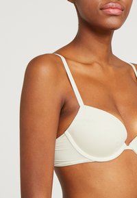 Light beige bra with molded cups, adjustable straps, lace trim along the top, and a smooth texture. Band features subtle branding.