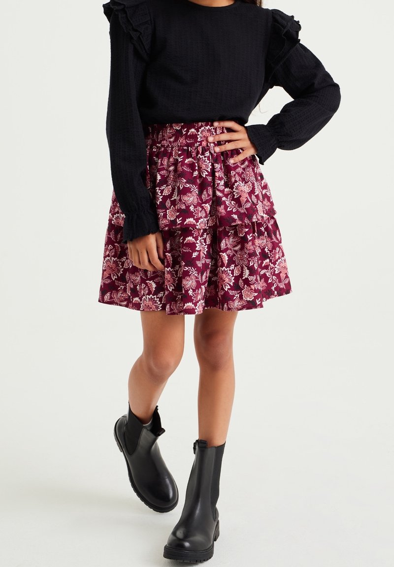 Fille portant un haut noir à manches longues avec des volants, une jupe portefeuille à volants à fleurs bordeaux, et des bottines noires, debout avec une main sur la hanche.