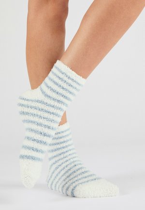 Beine mit flauschigen weißen Socken mit hellblauen horizontalen Streifen, ein Fuß über dem anderen auf einem schlichten hellen Hintergrund.