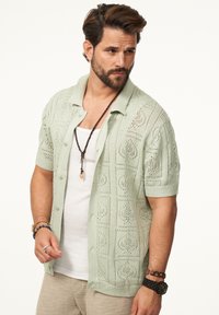 Hellgrünes, gestricktes kurzärmliges Shirt mit einem gemusterten, offenen Webdesign. Kombiniert mit einem weißen Tanktop und geschichteten Accessoires.
