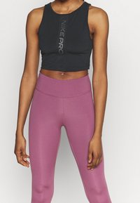 Osoba ubrana w czarny bezrękawnik crop top z napisem "NIKE PRO" i wysokim stanem fioletowe legginsy, stojąca na tle jednolitym.