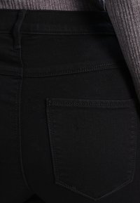 Black denim jeans feature a mid-rise waist, plain back pocket, with visible stitching and a smooth texture. The fabric appears durable and fitted.