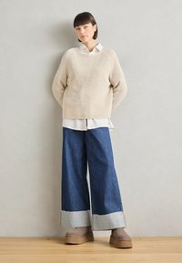 Pull côtelé beige porté sur une chemise blanche à col, assorti à un jean en denim bleu à jambes larges avec un ourlet effiloché et des accents gris clair. Chaussures à plateforme marron.