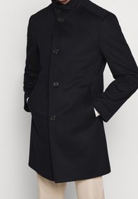 Cappotto in misto lana blu Navy con colletto alto, chiusura frontale a sei bottoni, tasche laterali e vestibilità strutturata. Tessuto liscio con dettagli minimi.