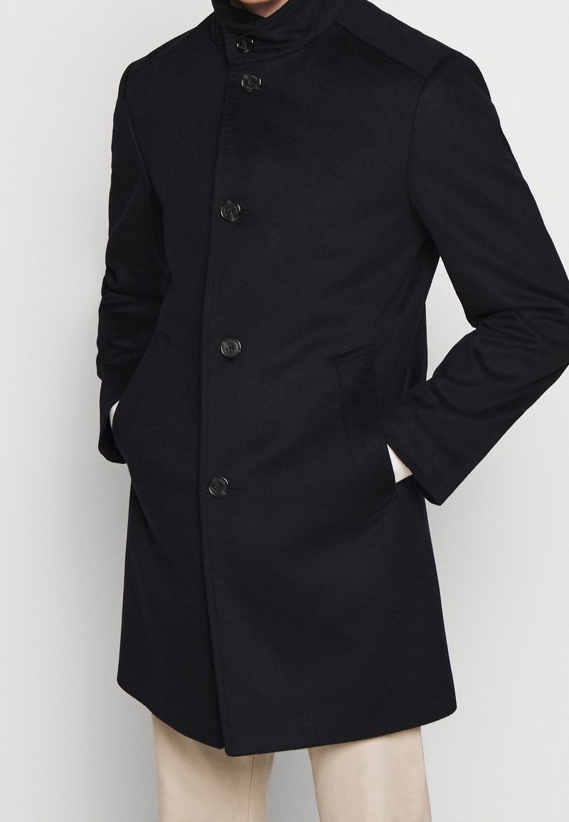 Cappotto in misto lana blu Navy con colletto alto, chiusura frontale a sei bottoni, tasche laterali e vestibilità strutturata. Tessuto liscio con dettagli minimi.