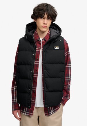 Giovane uomo con capelli ricci che indossa un gilet imbottito nero sopra una camicia a quadri rossa e pantaloni beige, in piedi davanti a uno sfondo bianco semplice.