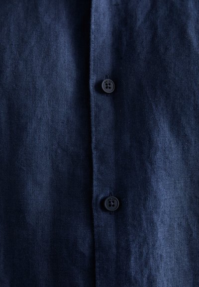 Camisa de lino azul marino con una superficie texturizada, que cuenta con dos botones negros y un cuello clásico. La tela parece ligera y transpirable.