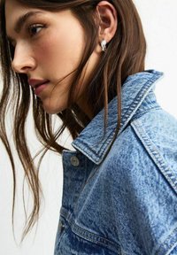 Giacca di denim azzurro chiaro con colletto classico, cuciture a vista e chiusura con bottoni. Il modello ha capelli lunghi e fluenti e indossa orecchini d'argento.