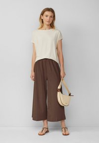 Vrouw staat tegen een eenvoudige muur en draagt een beige t-shirt met korte mouwen, brede bruine, cropped broek, sandalen met een luipaardprint, en houdt een crèmewitte, halvemaanvormige tas vast.