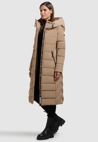 Dlouhý béžový prošívaný kabát s kapucí, zdobený cik-cak prošíváním, se zipovými bočními kapsami a štítkem s logem. Nošený s černými kozačkami a legínami.